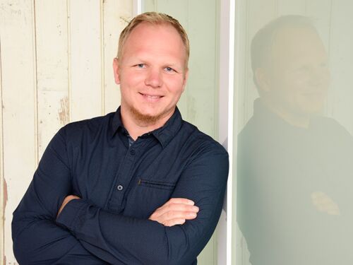 Auf dem Foto ist Tim Krenke. Er hat blond-rötliches Haar, einen kurzen Bart und lächelt. Er trägt ein dunkelblaues Hemd und lehnt an einer hellen Hauswand.